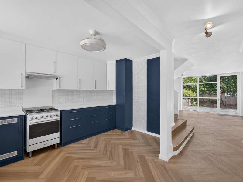 Modern open-plan kitchen featuring two-tone cabinetry, gas stove, and herringbone timber flooring, flowing into a bright living area with direct garden access.