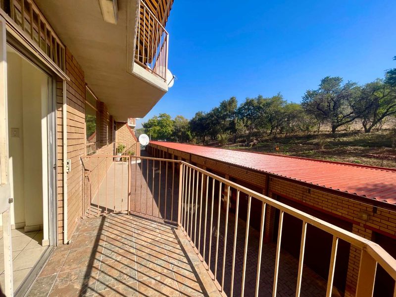 A spacious apartment balcony featuring tiled flooring and metal balustrades, overlooking the complex grounds and garage block.