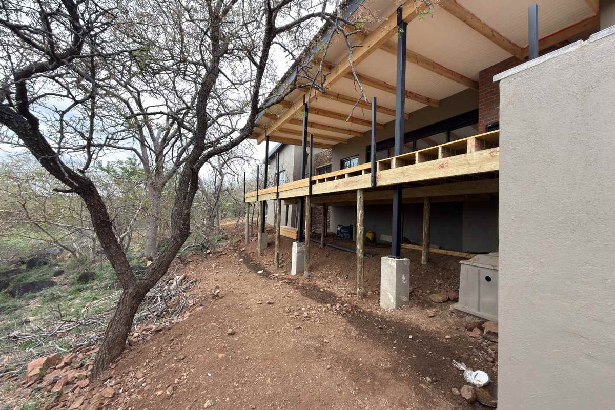Rear view of a house featuring an unfinished raised timber deck with exposed steel supports and timber rafters, set against a natural bushveld landscape.