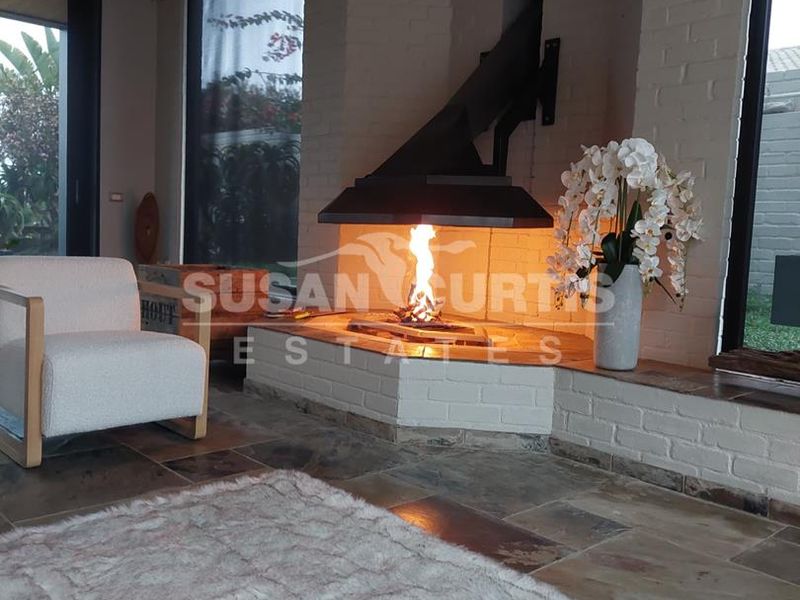 A cozy living room featuring a prominent corner fireplace with a metal flue, stone flooring, and large windows overlooking the garden.