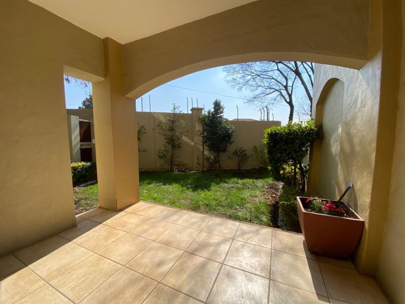 A covered patio area featuring tiled flooring and arched structural pillars, overlooking a private garden space enclosed by a perimeter wall with electric fencing.