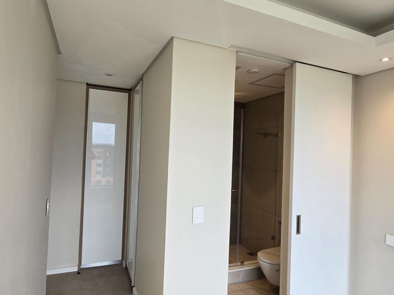 A modern, minimalist bedroom featuring neutral tones, recessed ceiling lighting, built-in storage, and a sliding door leading to an en-suite bathroom.