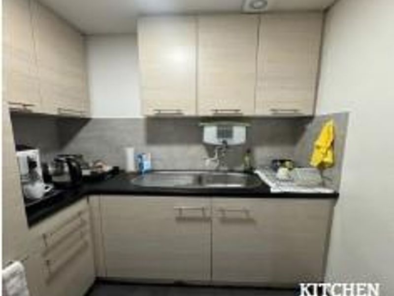 A compact office kitchenette featuring light wood-tone cabinetry, a stainless steel sink, and a tiled backsplash.