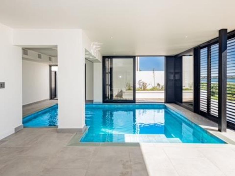 A modern indoor swimming pool area featuring large sliding glass doors, folding shutters, and sleek tiled flooring, offering a seamless transition to the outdoors.