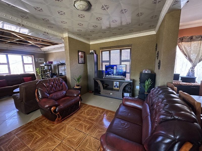 Living room featuring tiled flooring and decorative ceiling tiles, with an adjacent area showing exposed structural beams and unfinished ceiling work.