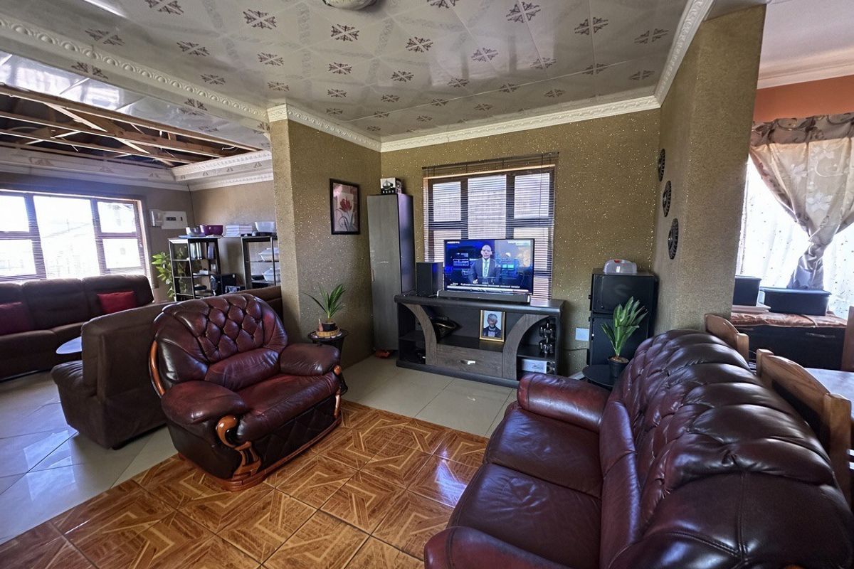 Living room featuring tiled flooring and decorative ceiling tiles, with an adjacent area showing exposed structural beams and unfinished ceiling work.