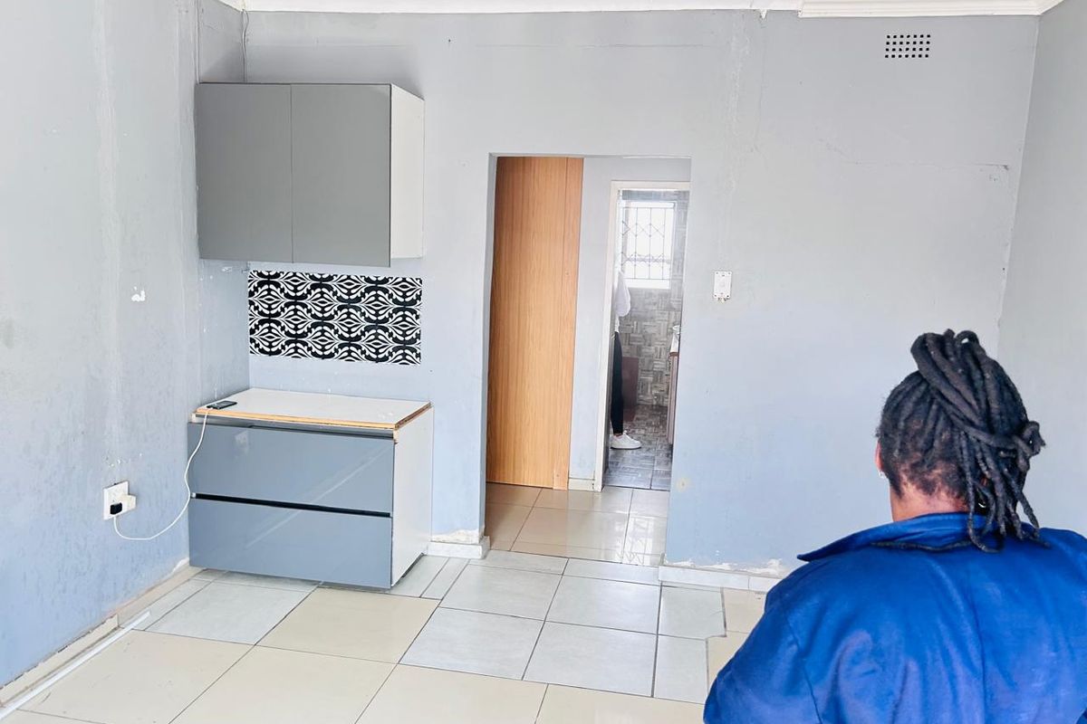 A small, empty kitchen area featuring grey built-in cabinetry, decorative backsplash tiles, and light-colored floor tiles, requiring cosmetic updates to improve its overall condition.