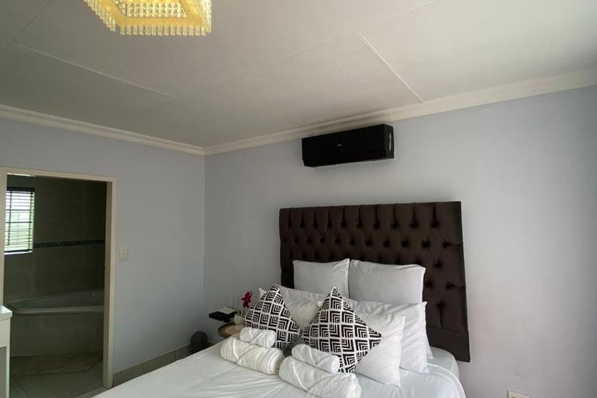 A well-maintained bedroom featuring tiled flooring, a modern crystal chandelier, and a wall-mounted air conditioning unit, with direct access to an en-suite bathroom.