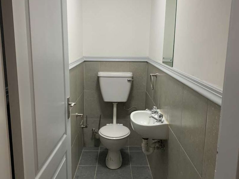 A compact industrial restroom featuring a wall-mounted basin, toilet, tiled lower walls, and a drop ceiling.