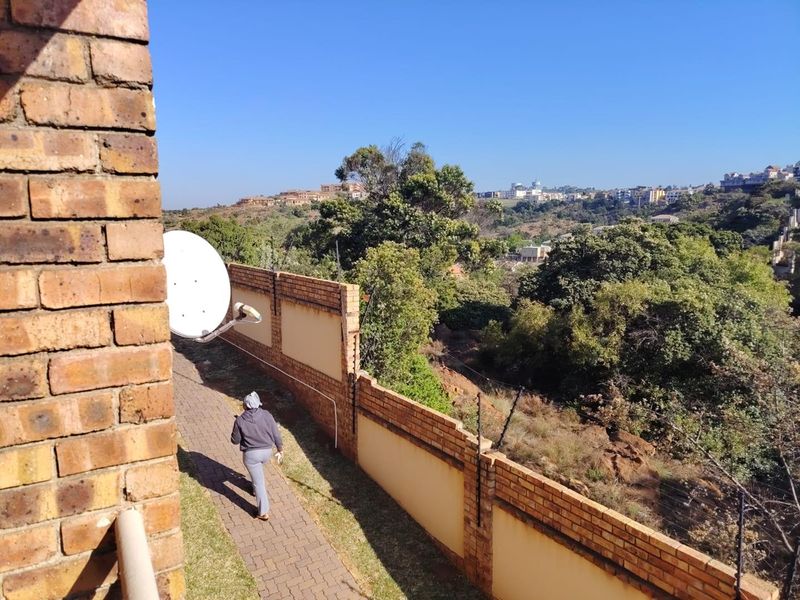 An elevated view from the property showing a paved walkway, brick boundary wall with electric fencing, and surrounding landscape.