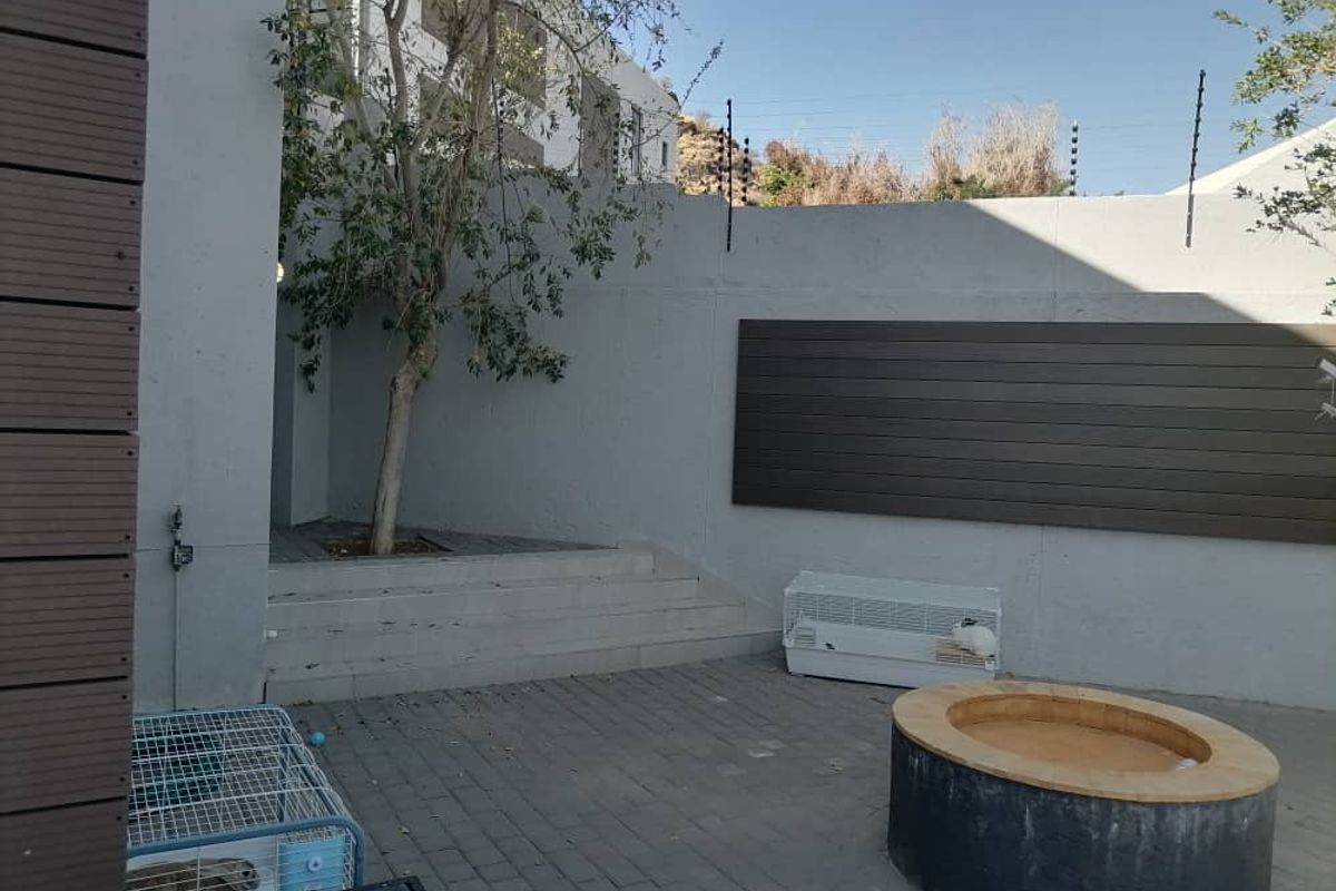A private rear courtyard featuring paved flooring, a built-in circular fire pit, raised garden bed with a tree, and perimeter security fencing.