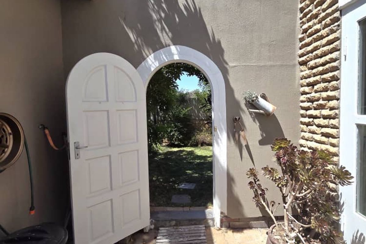A charming arched doorway leading from a paved courtyard into a lush rear garden, featuring rustic brickwork and a classic white panel door.