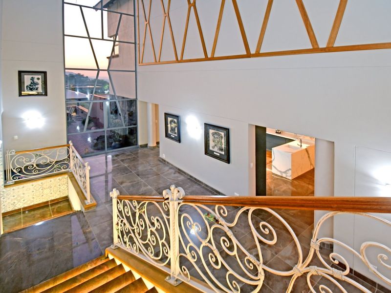 Spacious double-volume entrance hall featuring elegant wrought iron balustrades, large floor-to-ceiling windows, and polished tiled flooring with a view into the kitchen area.