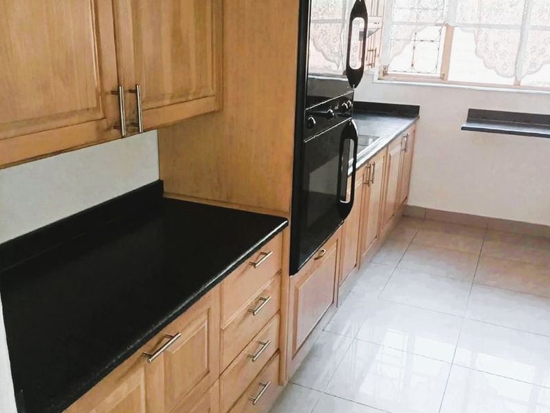 A clean, functional kitchen featuring light wood cabinetry, contrasting black countertops, built-in oven, and neutral tiled flooring with ample natural light.