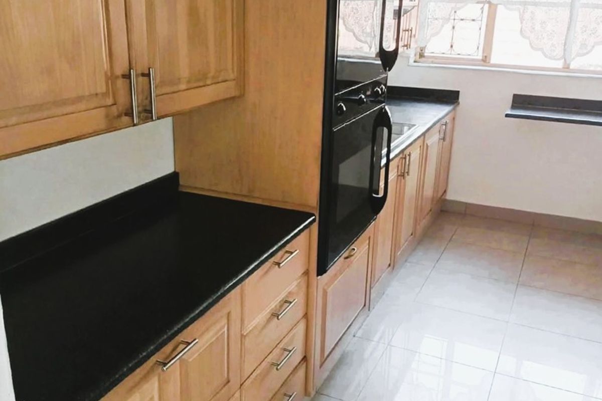 A clean, functional kitchen featuring light wood cabinetry, contrasting black countertops, built-in oven, and neutral tiled flooring with ample natural light.