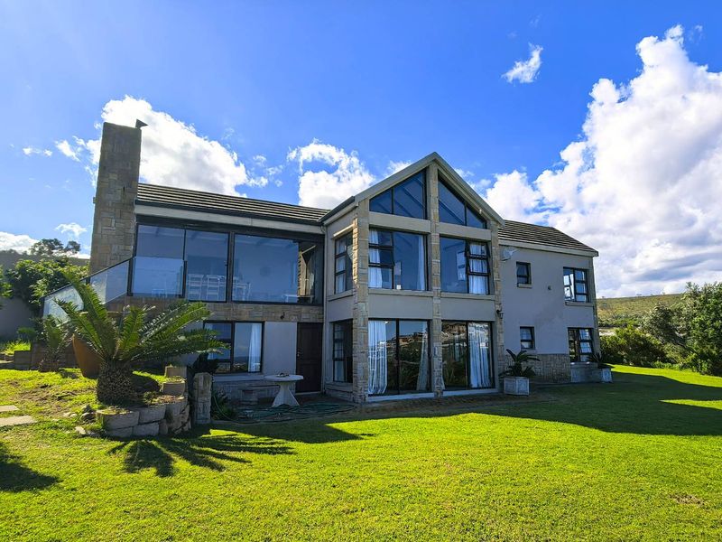 A spacious double-story residence featuring extensive glass windows, stone cladding, a prominent chimney, and a large balcony overlooking a well-maintained garden.