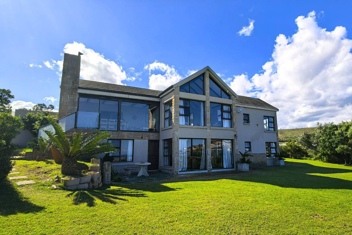 A spacious double-story residence featuring extensive glass windows, stone cladding, a prominent chimney, and a large balcony overlooking a well-maintained garden.
