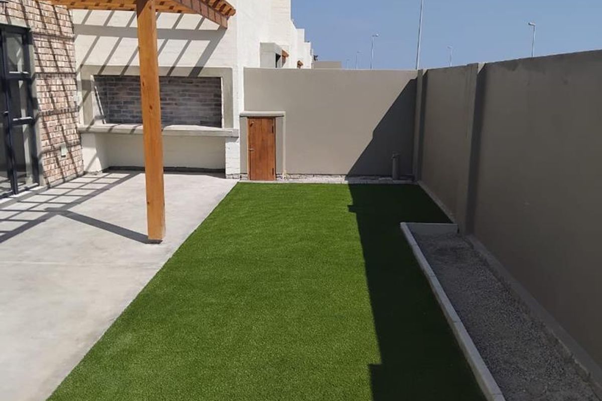 A well-maintained rear courtyard featuring a concrete patio with a wooden pergola, a built-in braai, and a neat artificial lawn area.
