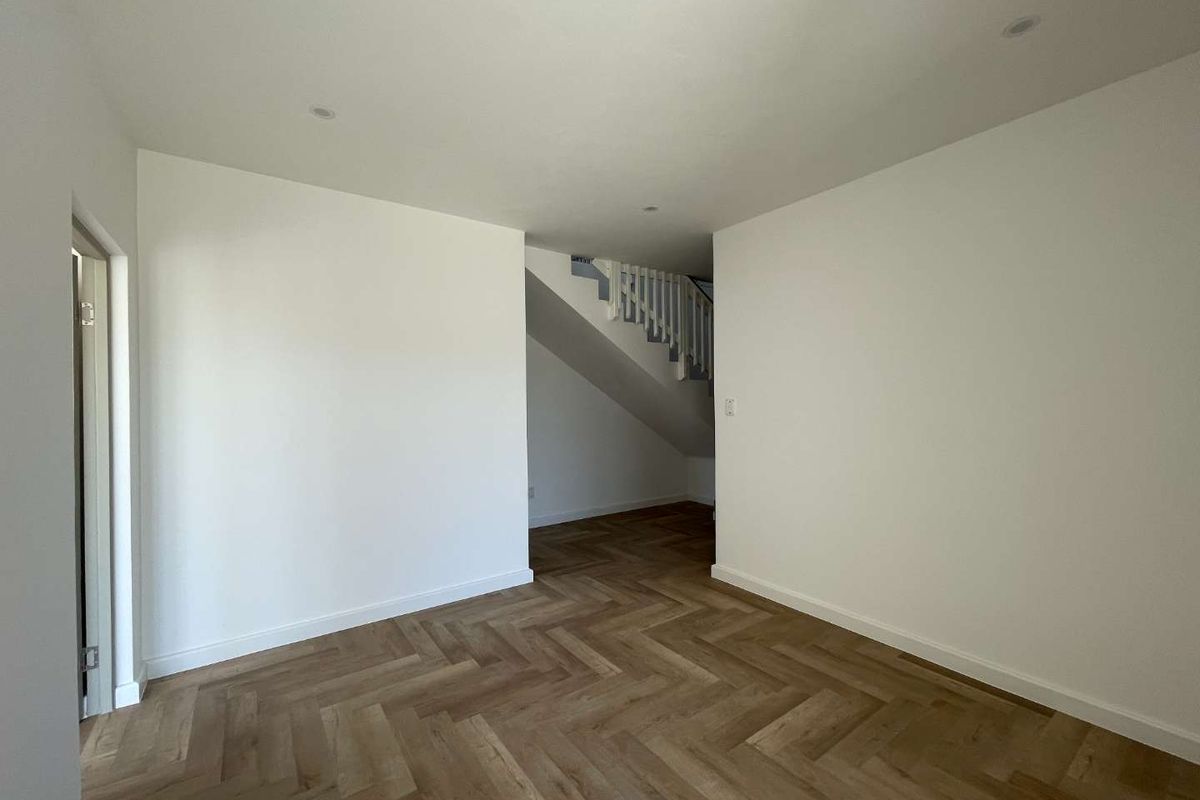 A bright, empty living area featuring modern herringbone wood-look flooring and a staircase with a white balustrade in the background.