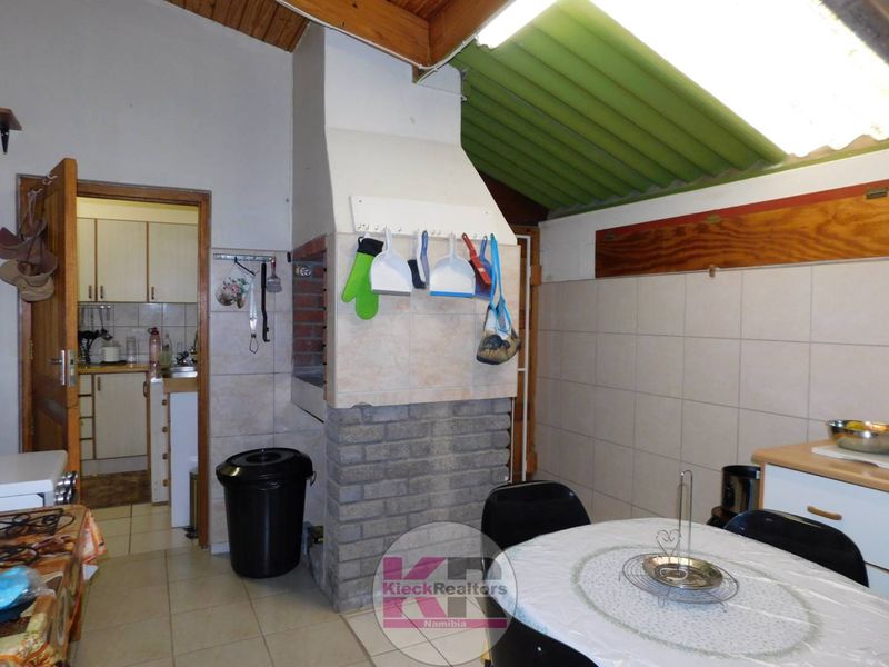 Open plan living area featuring a built-in braai, tiled flooring, and exposed timber ceiling, with an adjacent kitchen area visible.