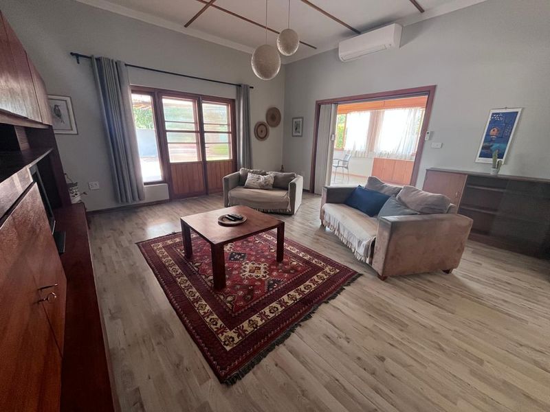 Spacious living room featuring light-toned laminate flooring, timber-framed glass doors leading to an outdoor area, and a wall-mounted split-system air conditioner.