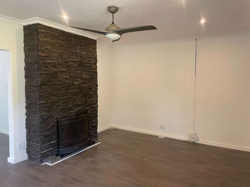 A spacious living room featuring a prominent stone-clad fireplace, laminate flooring, and a ceiling fan, offering a neutral canvas for interior design.