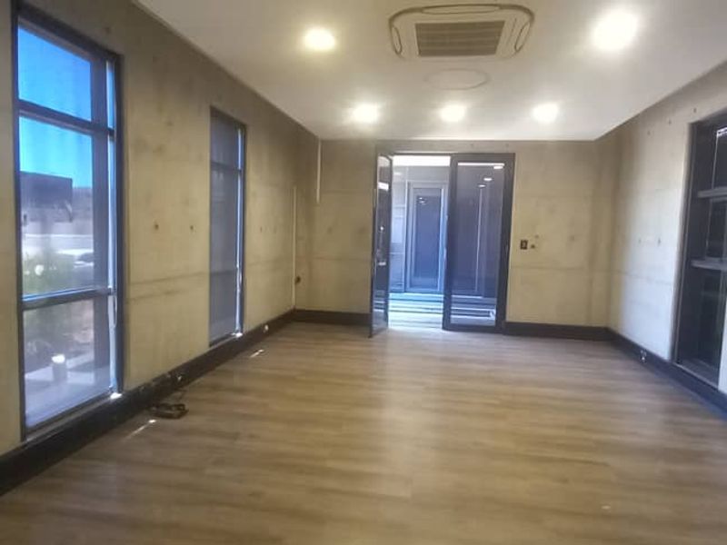 A modern, empty office space featuring exposed concrete walls, laminate flooring, recessed lighting, and large windows providing ample natural light.