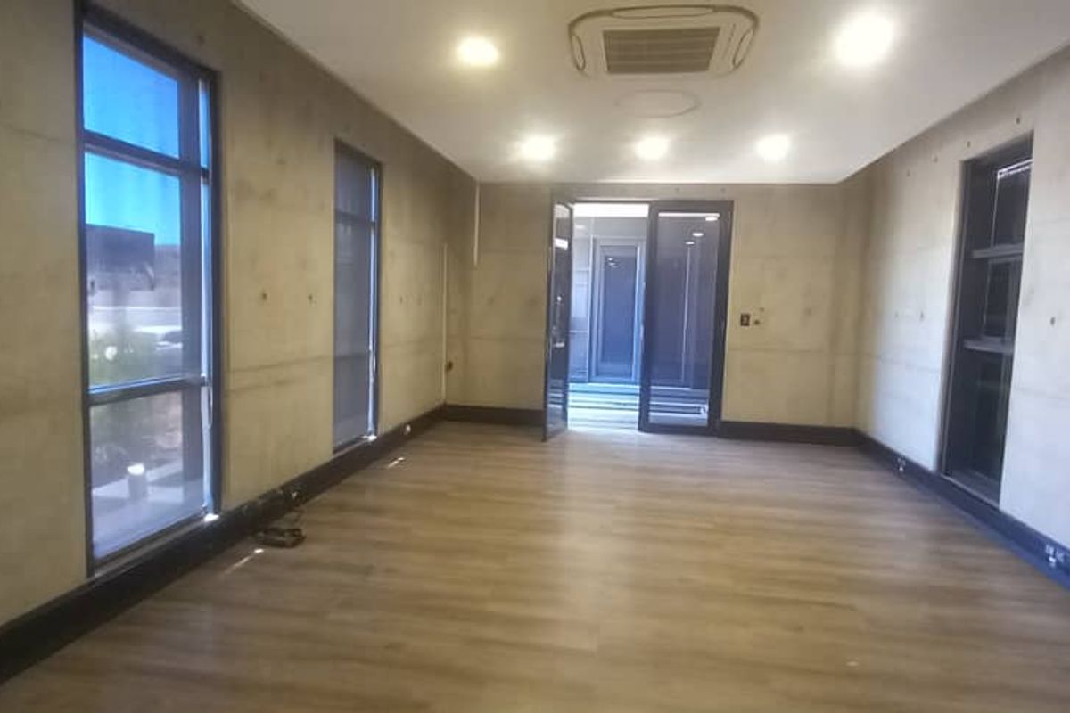 A modern, empty office space featuring exposed concrete walls, laminate flooring, recessed lighting, and large windows providing ample natural light.