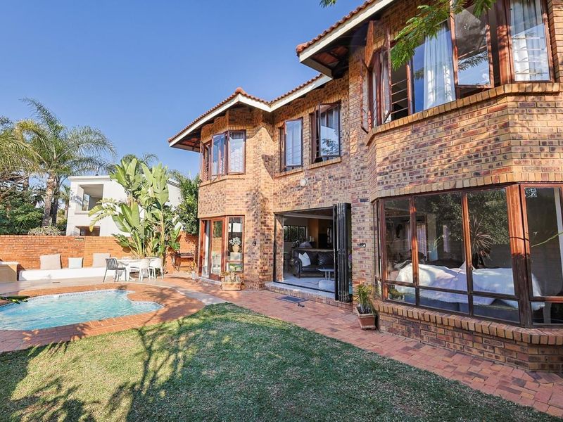 Rear view of a double-story brick house featuring a swimming pool, paved patio area, and large windows with bi-fold doors leading to the interior.
