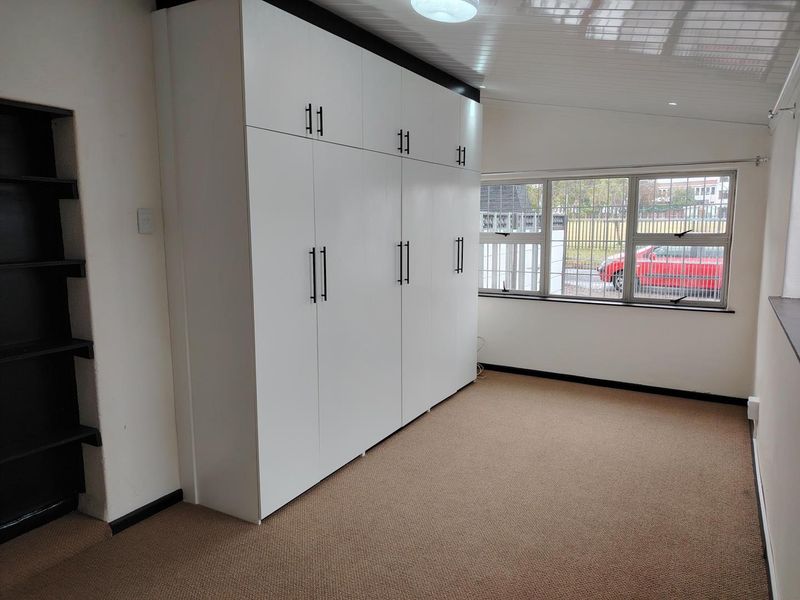 A bright, empty bedroom featuring extensive built-in white cabinetry, built-in shelving, carpeted flooring, and large windows with security bars.