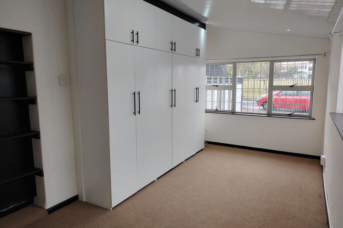 A bright, empty bedroom featuring extensive built-in white cabinetry, built-in shelving, carpeted flooring, and large windows with security bars.