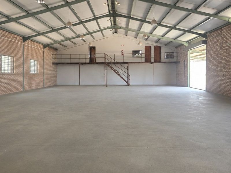 Spacious industrial warehouse featuring high ceilings with exposed steel trusses, concrete flooring, brick walls, and a functional mezzanine level with staircase access.