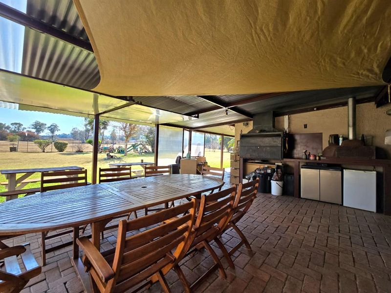 Covered outdoor patio area featuring a built-in braai, paved flooring, and a shade sail, overlooking the expansive garden grounds.