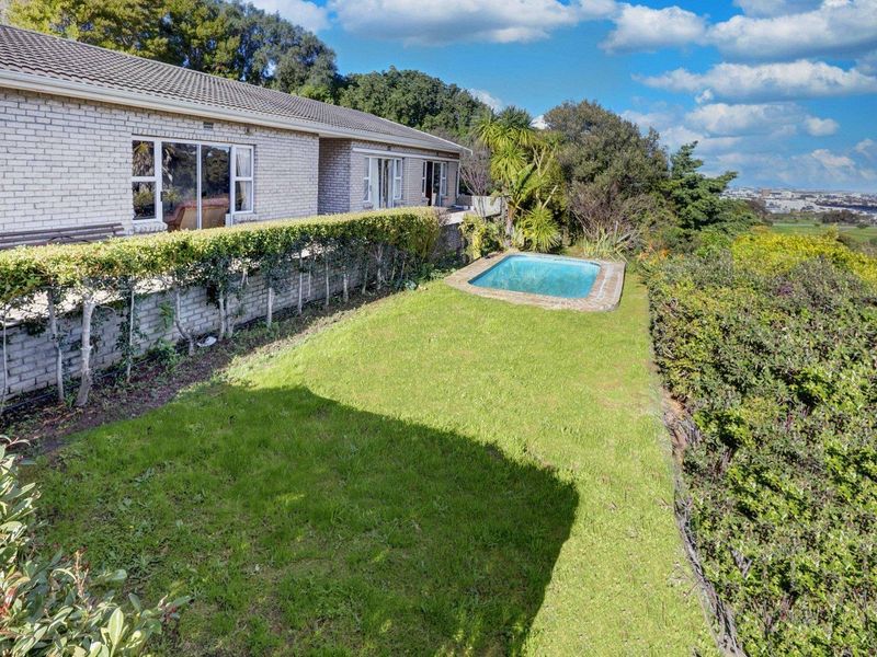 Rear exterior view featuring a brick house, a small swimming pool, a manicured lawn, and established garden hedges with distant landscape views.