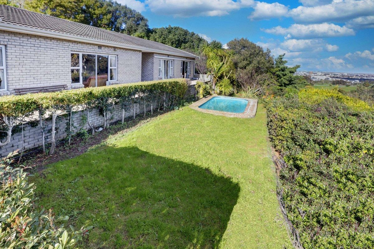 Rear exterior view featuring a brick house, a small swimming pool, a manicured lawn, and established garden hedges with distant landscape views.