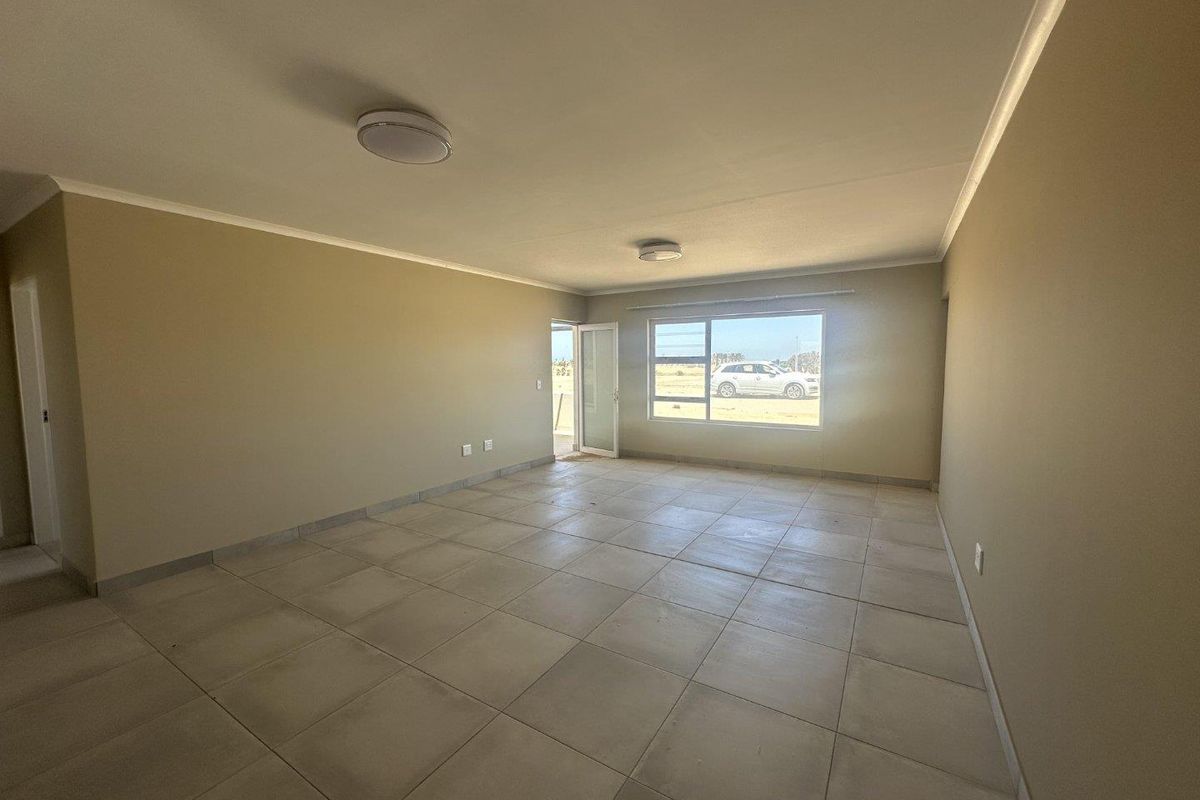A spacious, empty living area featuring neutral tiled flooring, large windows for natural light, and a glass door leading to the exterior.