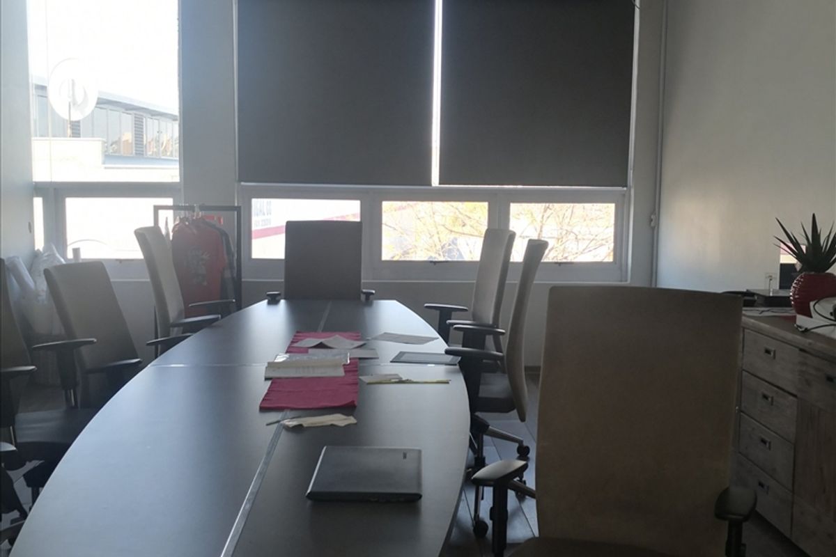 A functional office space featuring large windows with roller blinds, a suspended ceiling, and laminate flooring, suitable for a professional work environment.