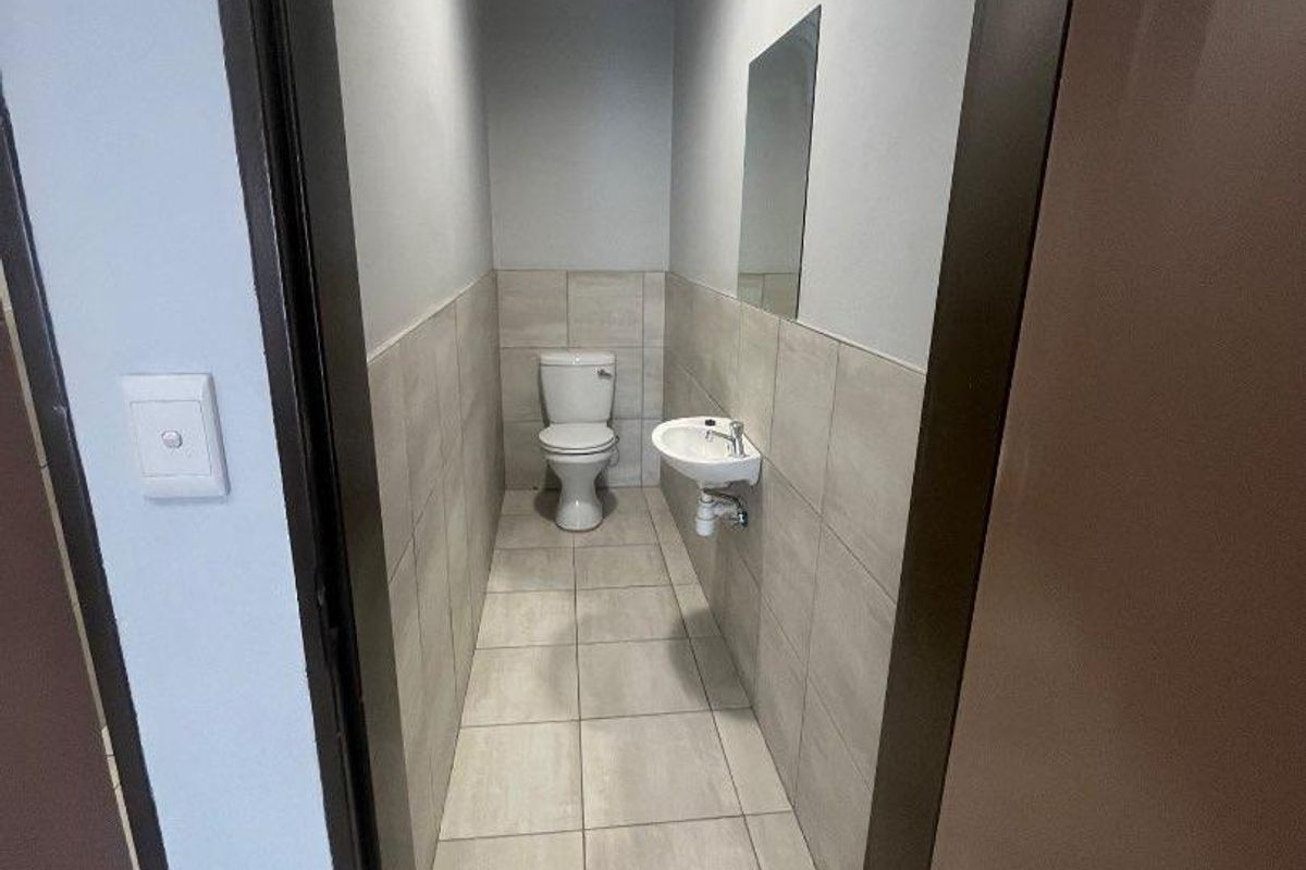 A clean, functional restroom featuring tiled flooring, wall-mounted basin, and toilet, finished with neutral wall tiles and a mirror.