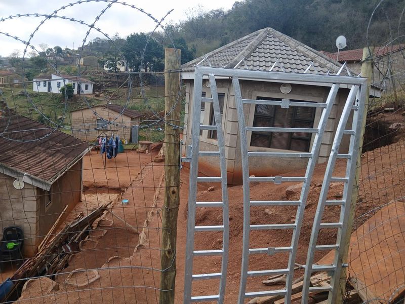 Exterior view of a residential property featuring a metal security gate, razor wire fencing, and an unlandscaped dirt yard area.