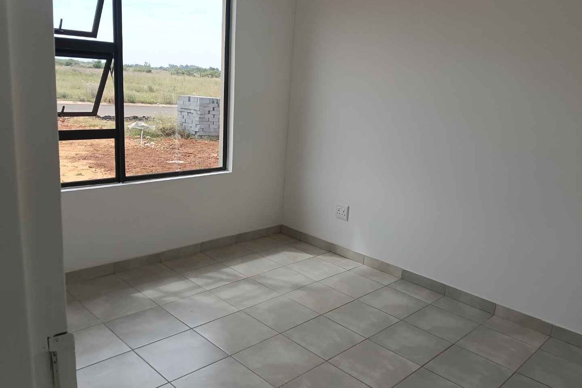 A bright, empty bedroom featuring neutral floor tiles, white walls, and a large window with black aluminum frames overlooking the exterior.
