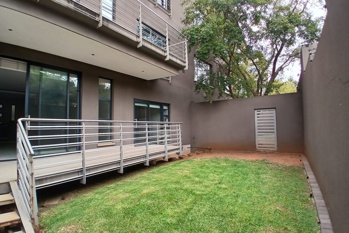Rear exterior view of the apartment building featuring a private garden, lawn area, and a metal balcony structure with sliding door access.