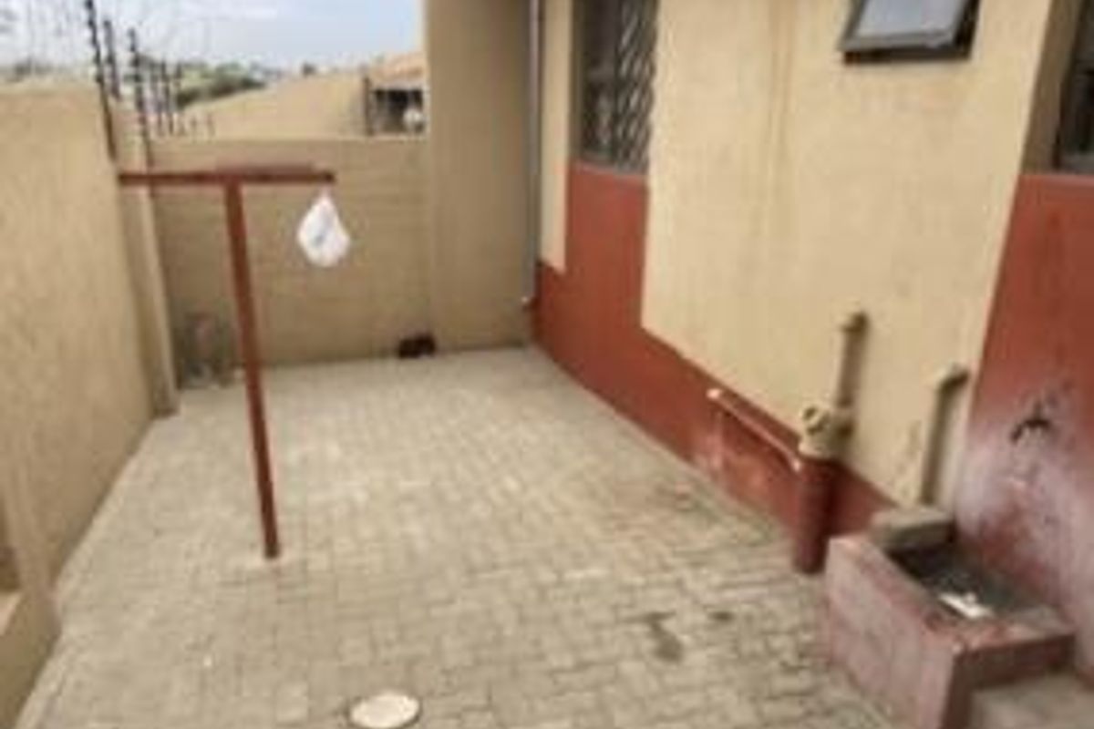 A paved rear courtyard area featuring a fixed washing line, external water tap, and security fencing, showing signs of wear and weathered exterior paint.