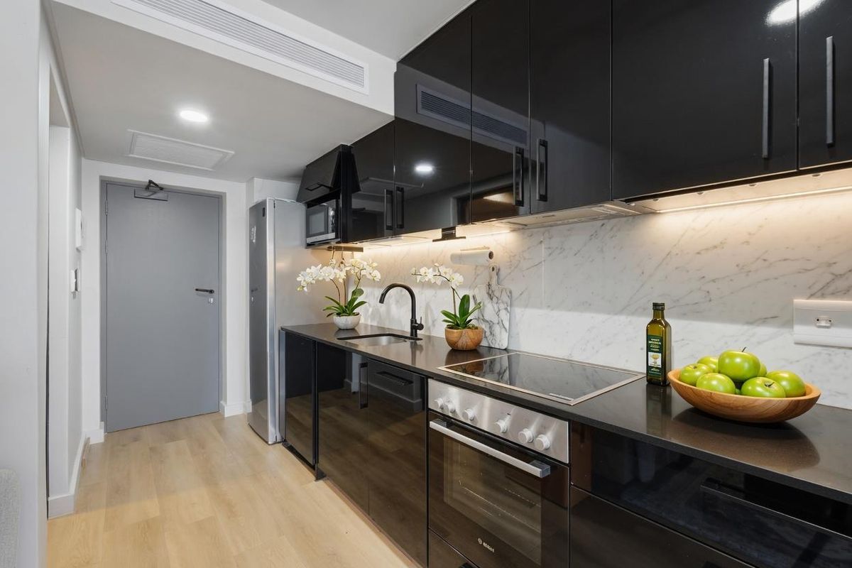 Modern kitchen featuring sleek black high-gloss cabinetry, marble-look splashback, stone countertops, and integrated appliances, complemented by recessed lighting and light timber-look flooring.