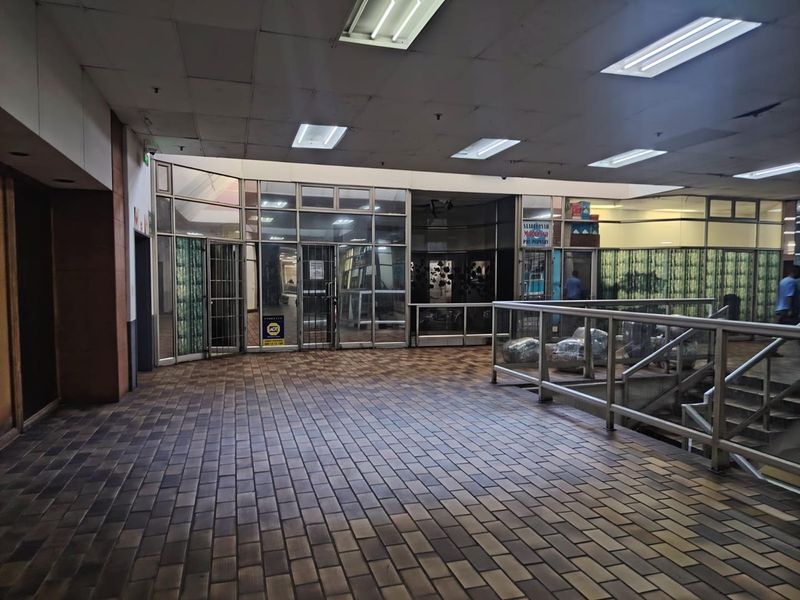 A vacant commercial hallway featuring tiled flooring, glass storefront partitions, and suspended ceiling lighting, showing signs of wear and maintenance requirements.