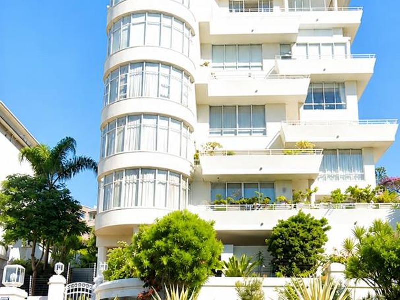 A striking multi-story penthouse building featuring a prominent curved glass tower, tiered balconies, and well-maintained landscaped gardens with a stone retaining wall.