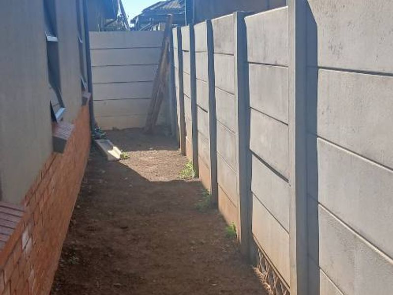 A narrow side passage featuring a precast concrete boundary wall, a metal carport structure, and an unlandscaped dirt ground surface.