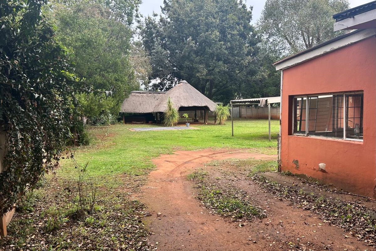 A view of the rear garden featuring a thatched lapa, large mature trees, and the side elevation of the house with a dirt driveway.