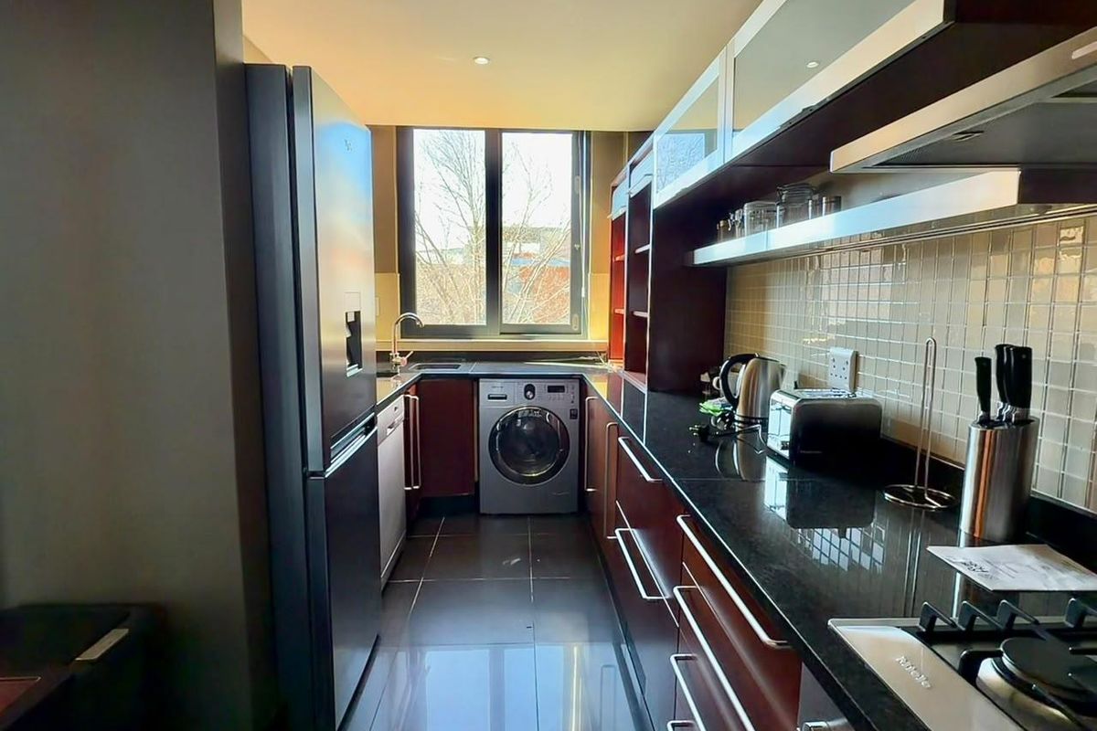 A modern galley-style kitchen featuring dark cabinetry, granite countertops, integrated appliances, and tiled flooring with natural light from a large window.