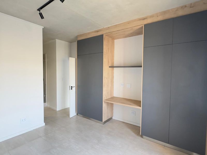 Modern bedroom featuring sleek built-in cupboards with an integrated desk area, neutral tiled flooring, and contemporary track lighting.
