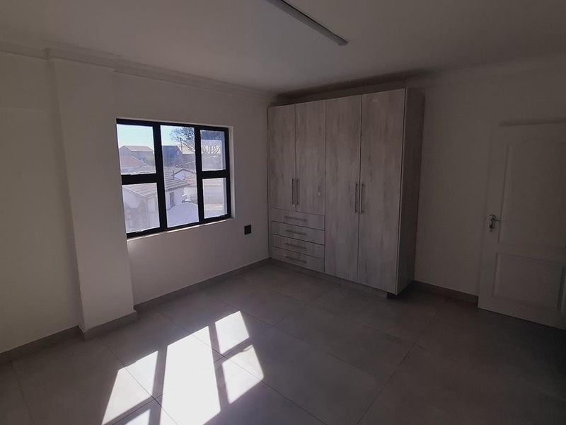 A bright, empty bedroom featuring neutral tiled flooring, built-in cupboards, and a modern aluminium window frame.